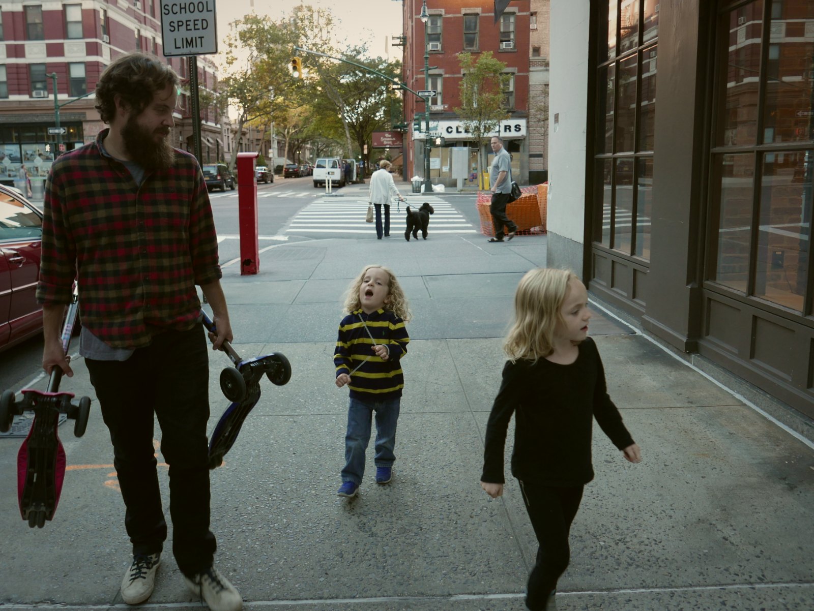 A dad walks his two young children along a city sidewalk on the way to school, carrying their scooters.