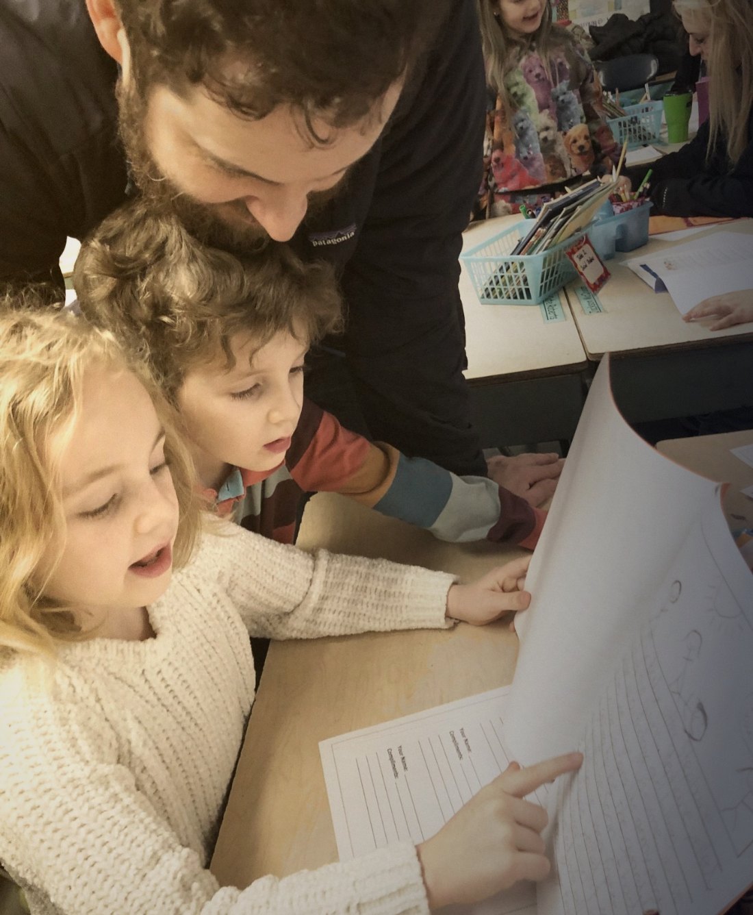 A dad leans in beside his two children at a classroom desk during a school visit, watching them work.