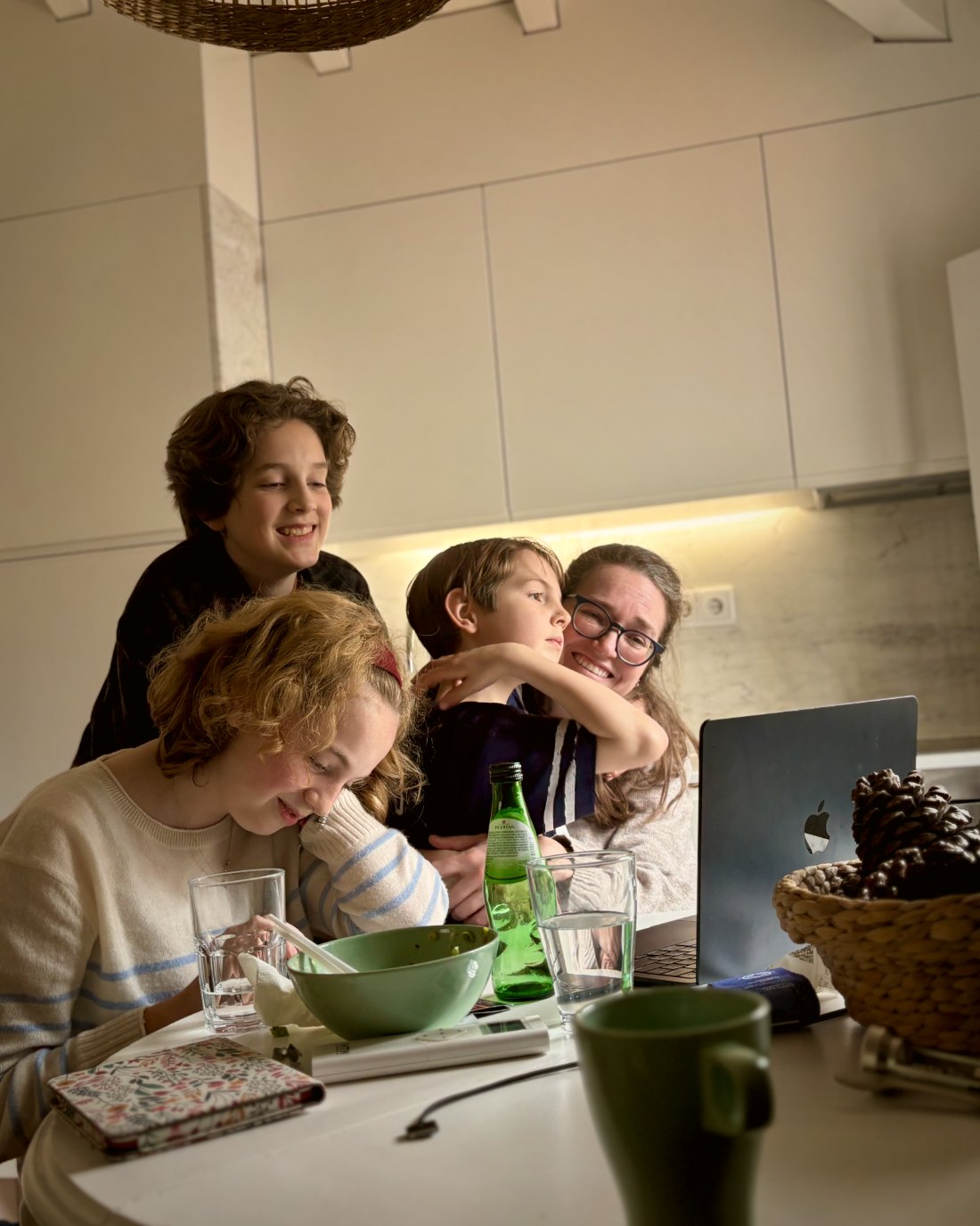 A mother and three children gather around a laptop at the kitchen table, smiling during a video call.