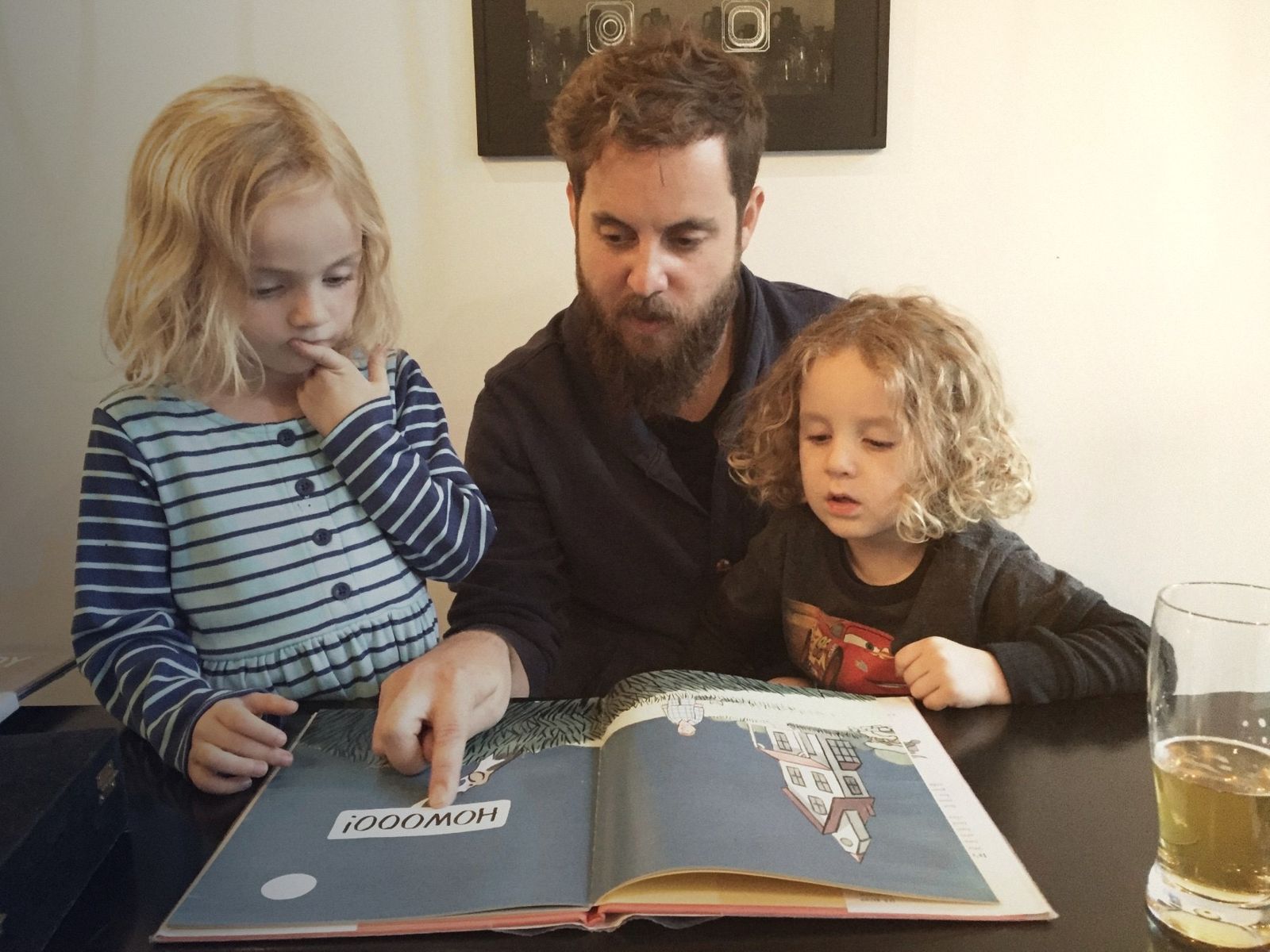 A dad reading a picture book at the table with his two young children, all three looking at the page.