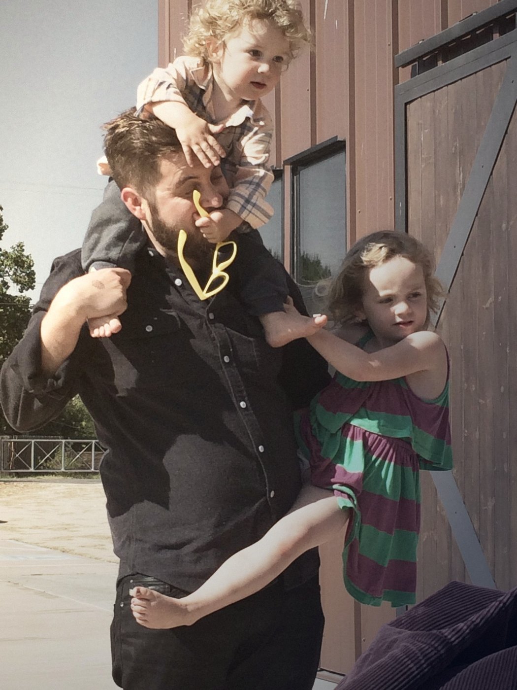 A dad carries one child on his shoulders while another climbs his side, late-afternoon sun on a wooden barn wall.